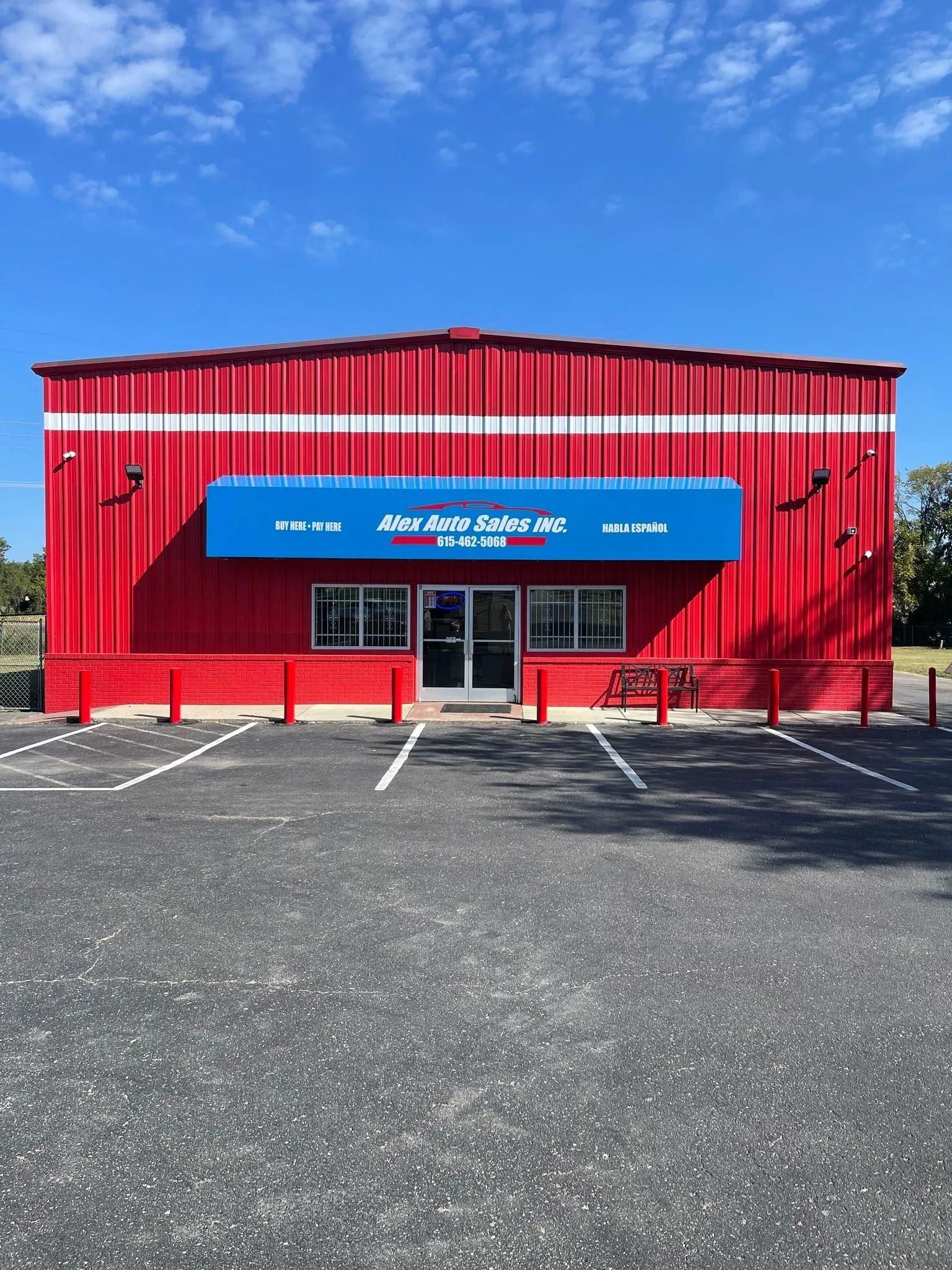A red metal building with a blue awning and glass front doors, situated in a paved parking lot under a sunny blue sky.