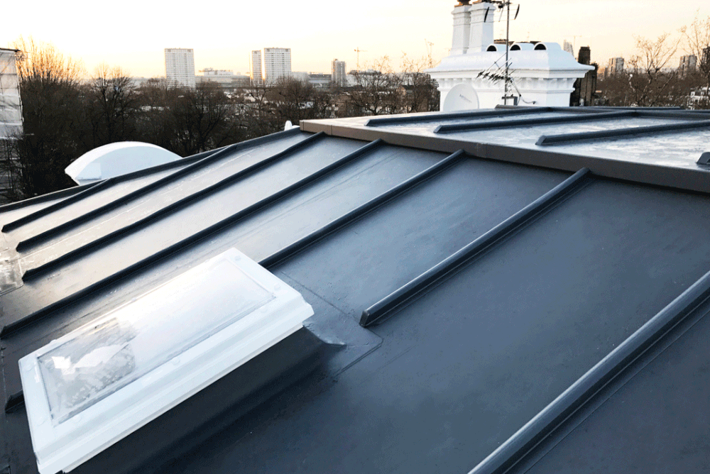 A dark gray standing seam metal roof with a white skylight, viewed from above at dusk with a city skyline in the distance.
