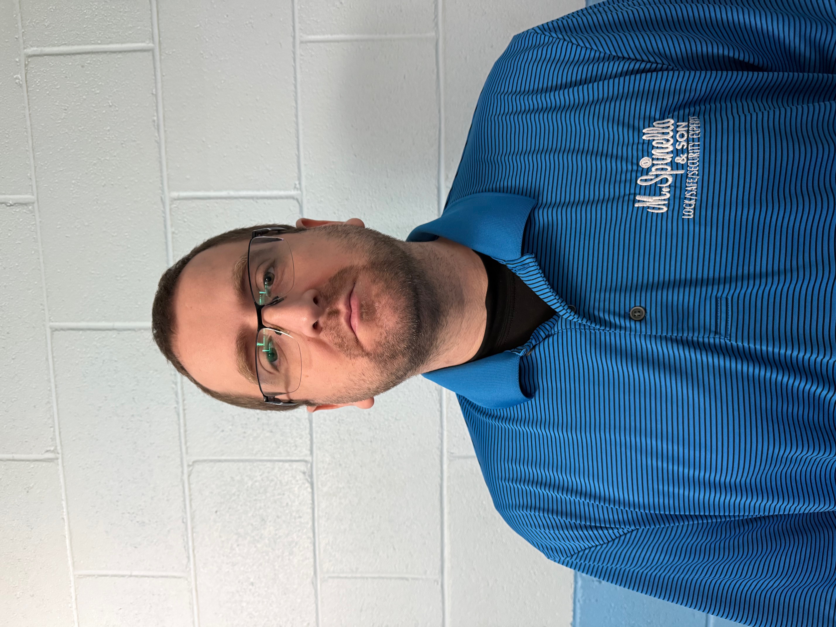 Man with glasses in a blue shirt stands against a white brick wall.