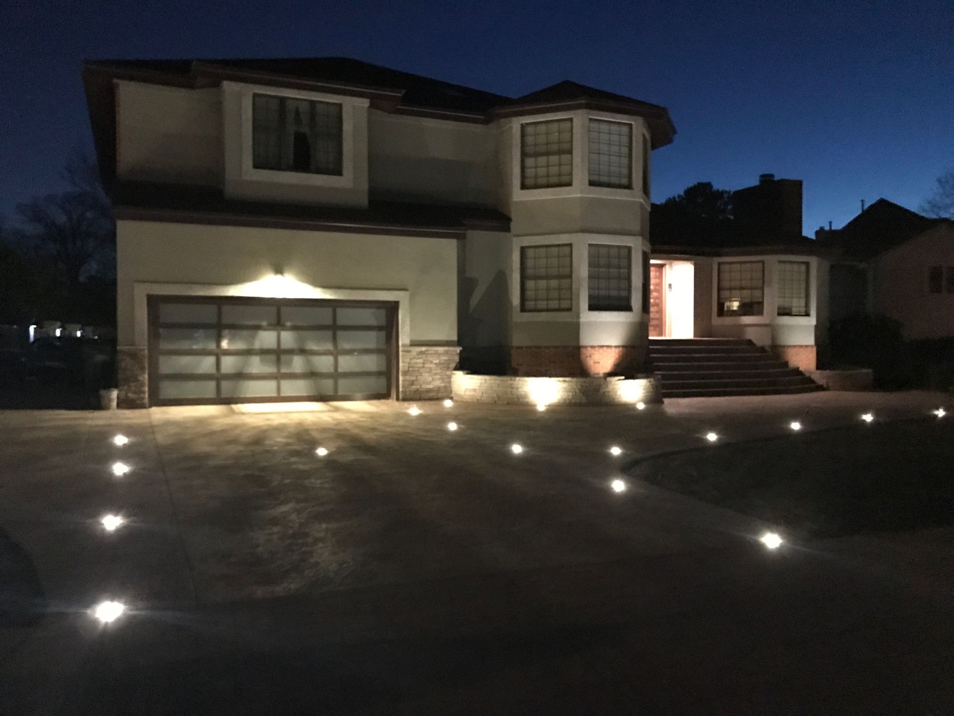 Two-story suburban house at night with lit driveway and steps, garage on left, illuminated entry on right