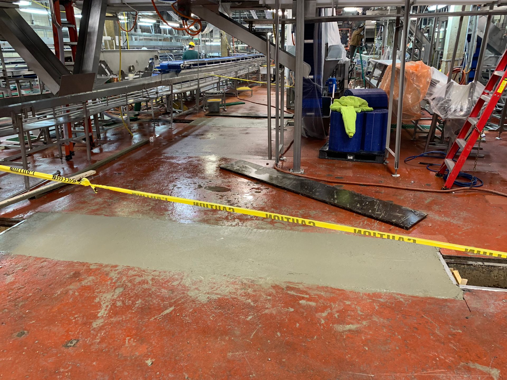 Factory floor with wet red concrete, caution tape, and workers near machinery under repair