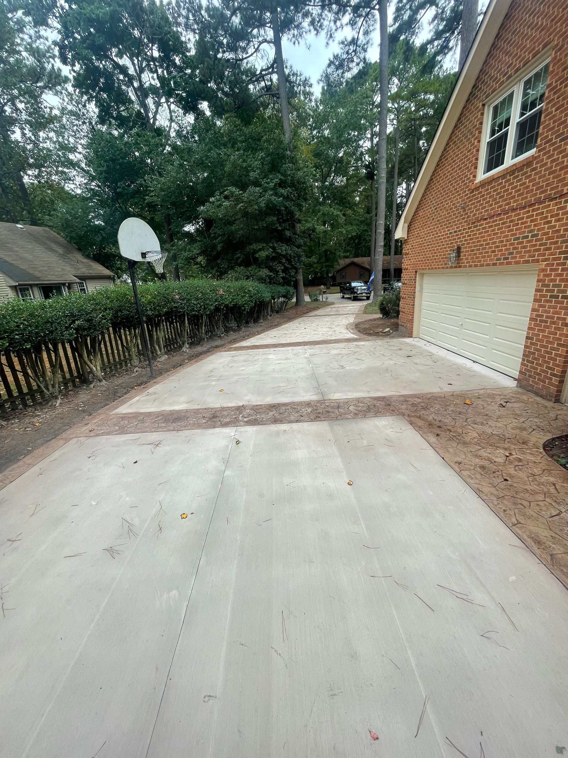 Freshly poured concrete driveway beside a brick house and garage, with a basketball hoop and trees along the side
