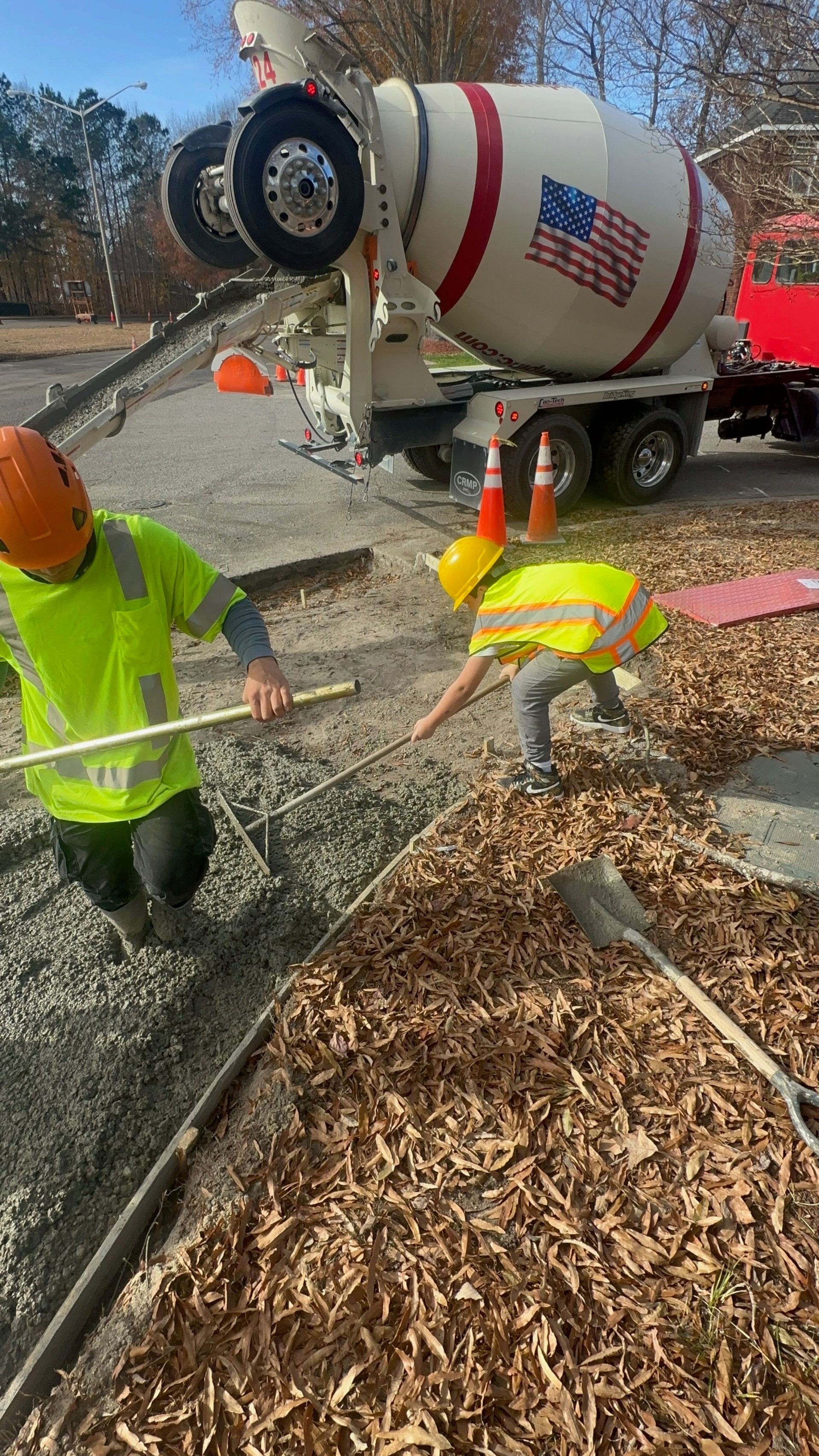 Road crew pouring concrete near a cement truck beside a curb and wood chips.