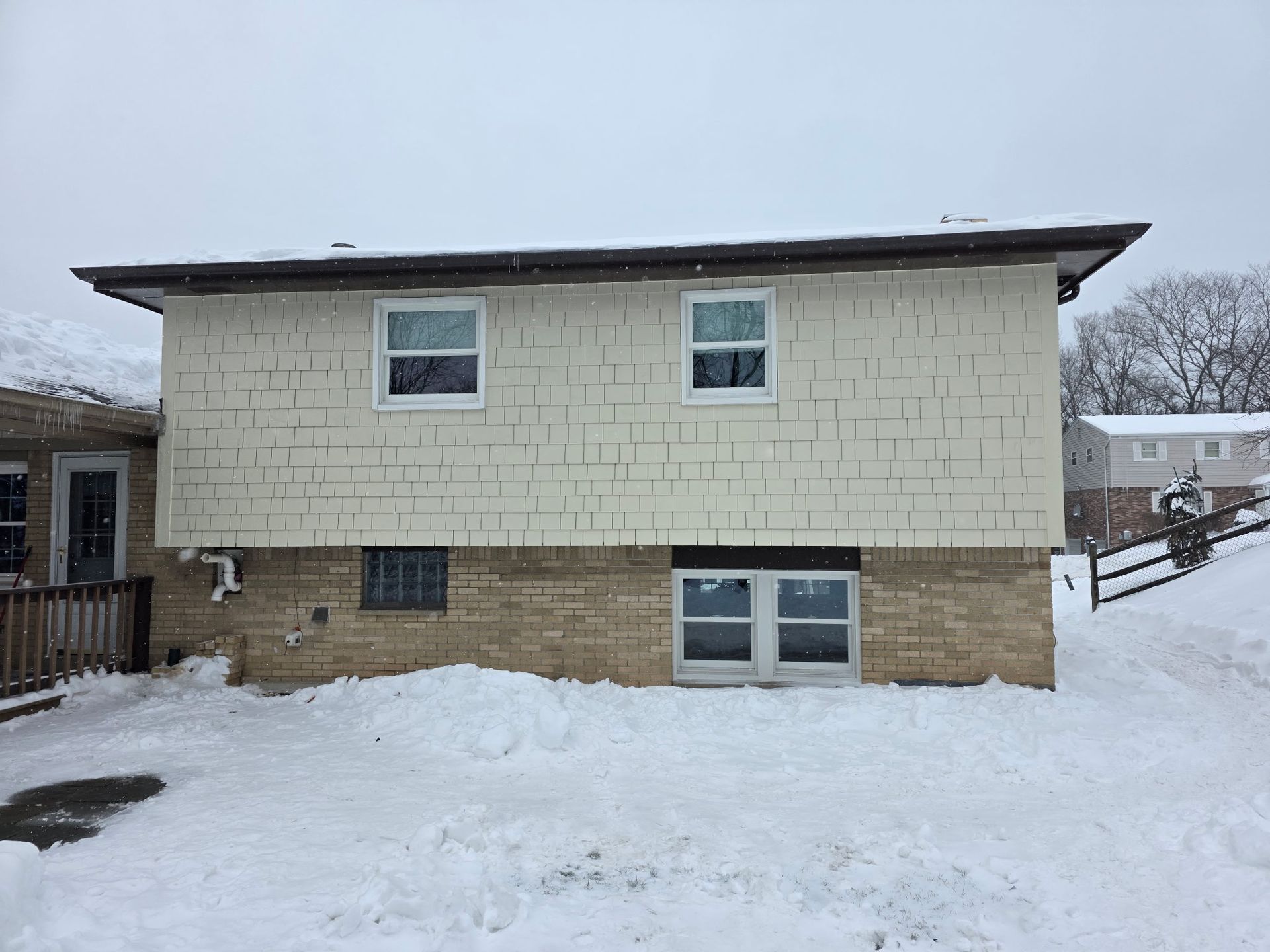 Two-story house with cream-colored siding and a tan brick foundation, covered in snow on a winter day.