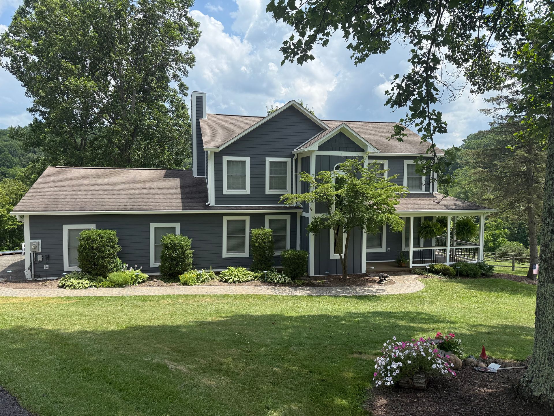 A two-story, dark blue home with a grey roof and front porch, surrounded by green lawns, trees, and small garden beds.