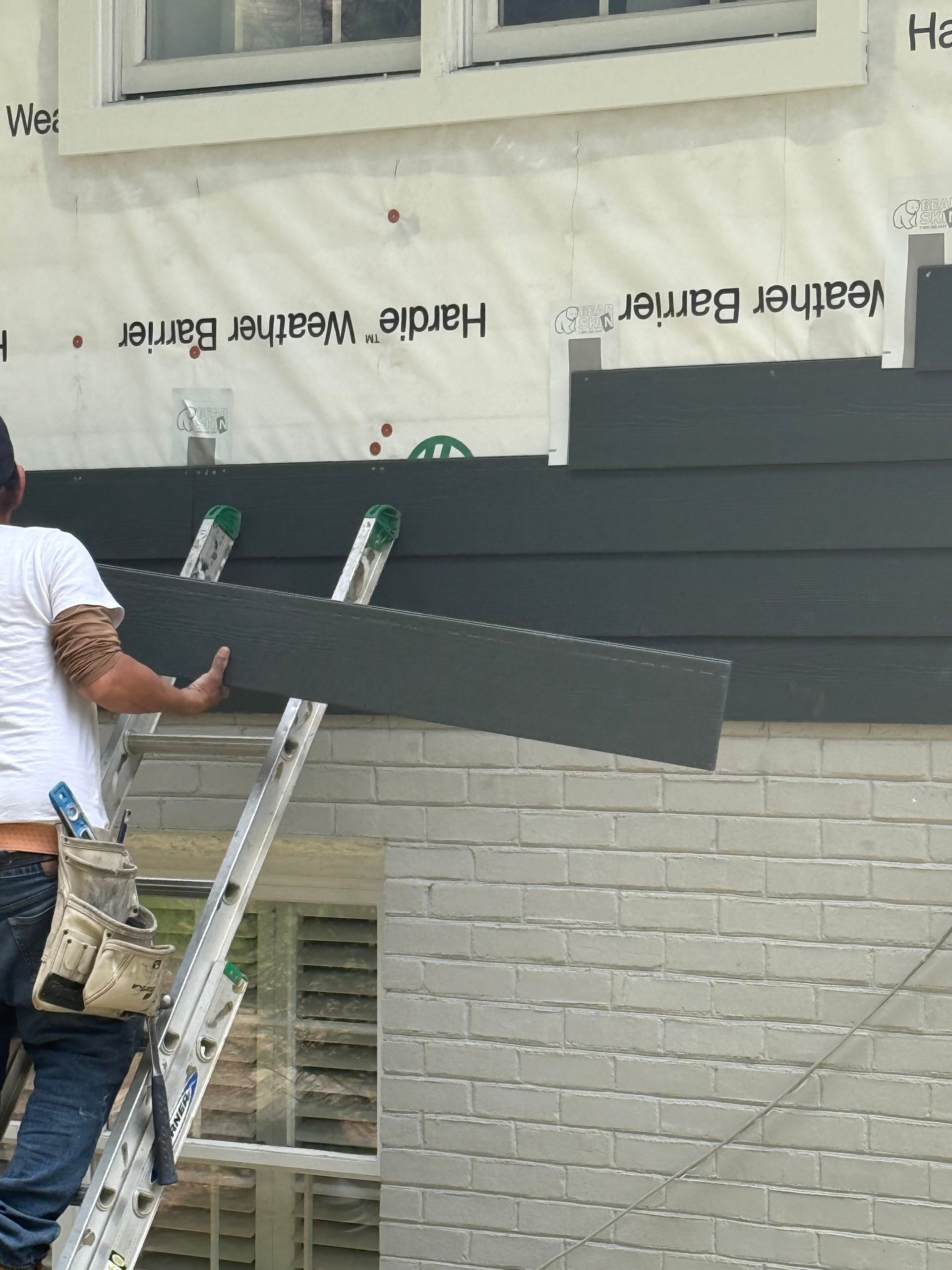 A construction worker on a ladder installs dark gray horizontal siding panels onto the side of a white brick building.
