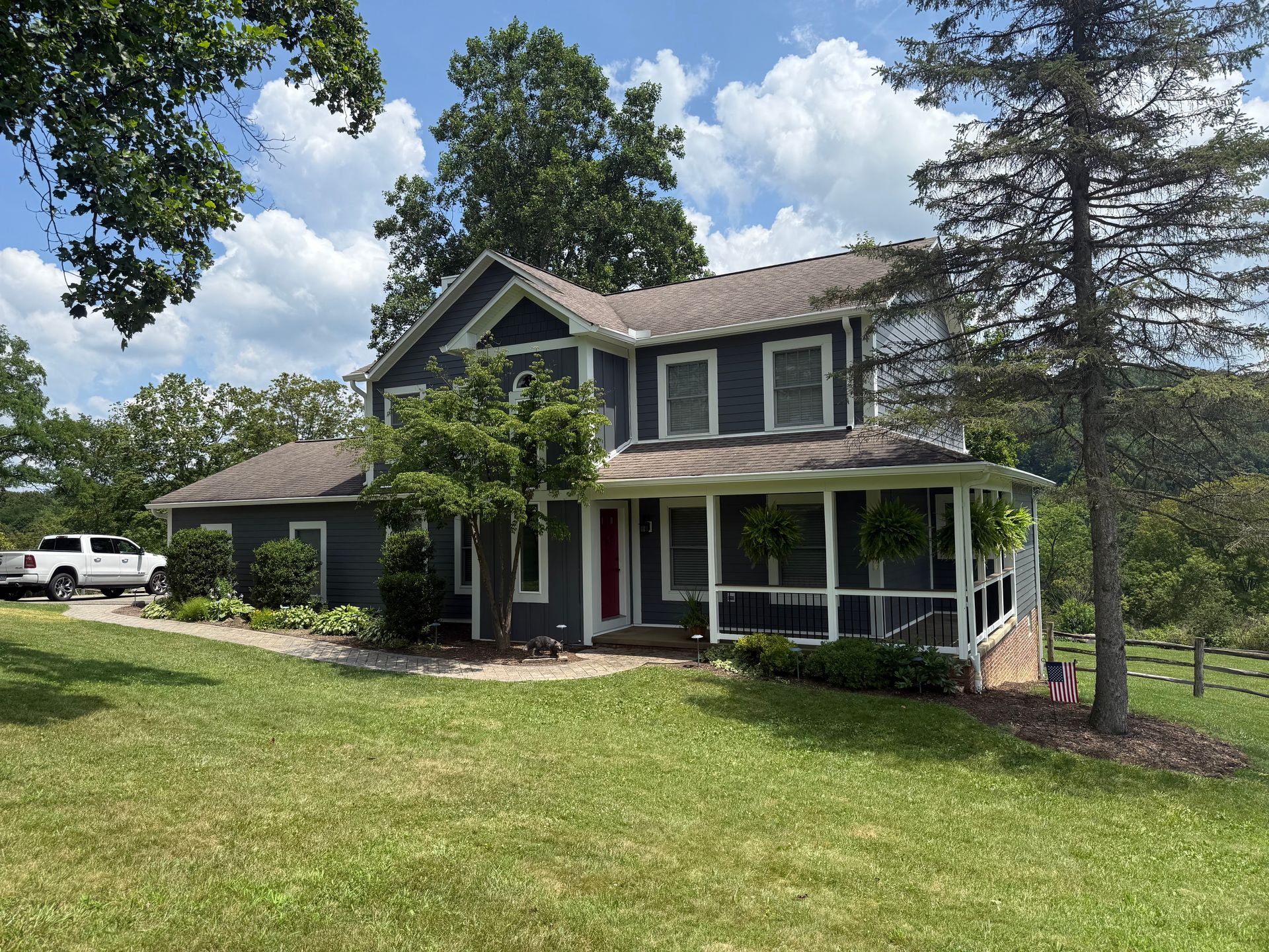 A two-story dark blue farmhouse with a white wrap-around porch, set on a grassy lawn with trees and a white pickup truck.