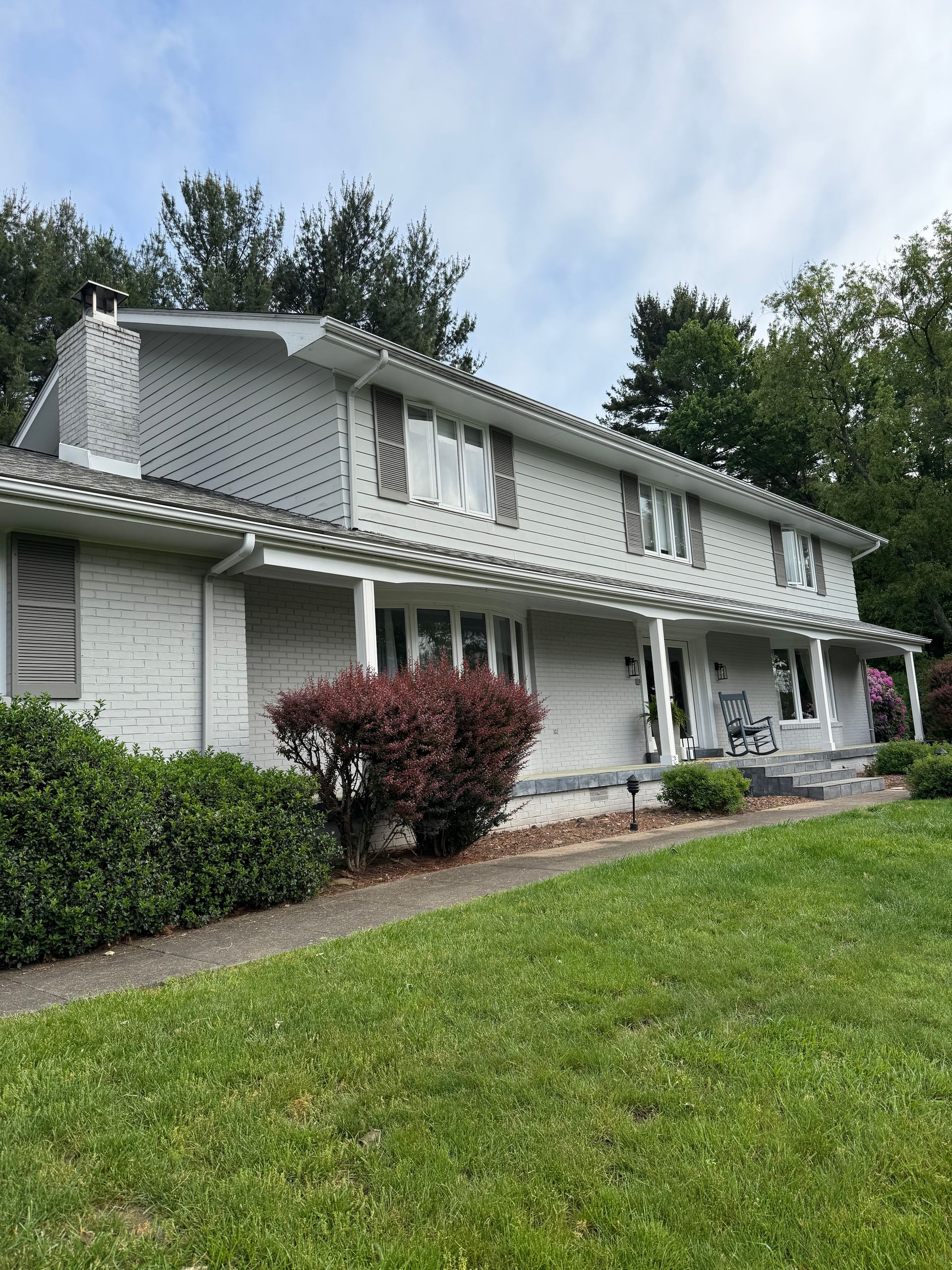 A two-story, white-painted brick house with a front porch, large windows, and a chimney, set behind a green lawn.