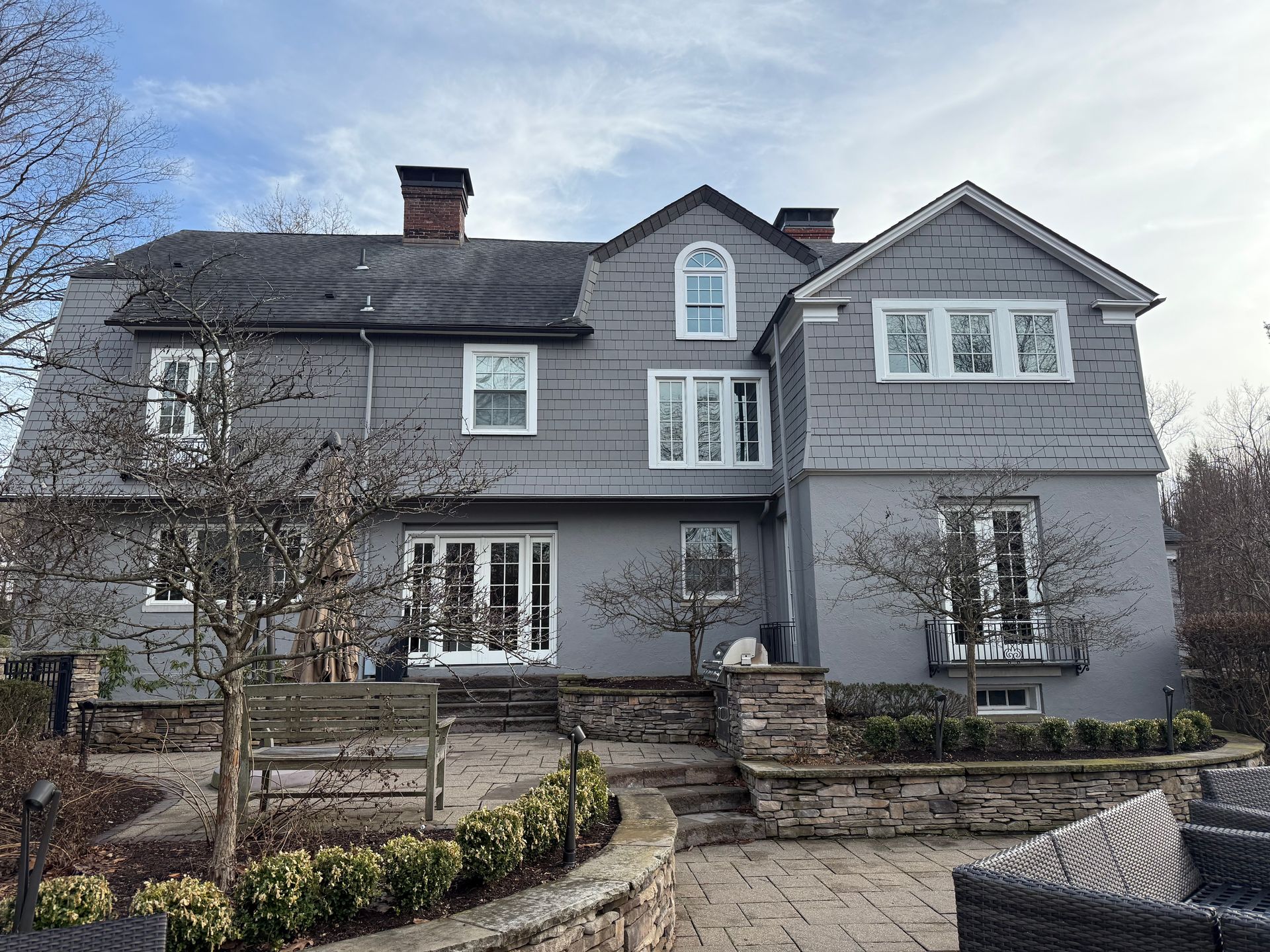 A large, multi-story gray house with a stone patio and retaining wall in the front yard.
