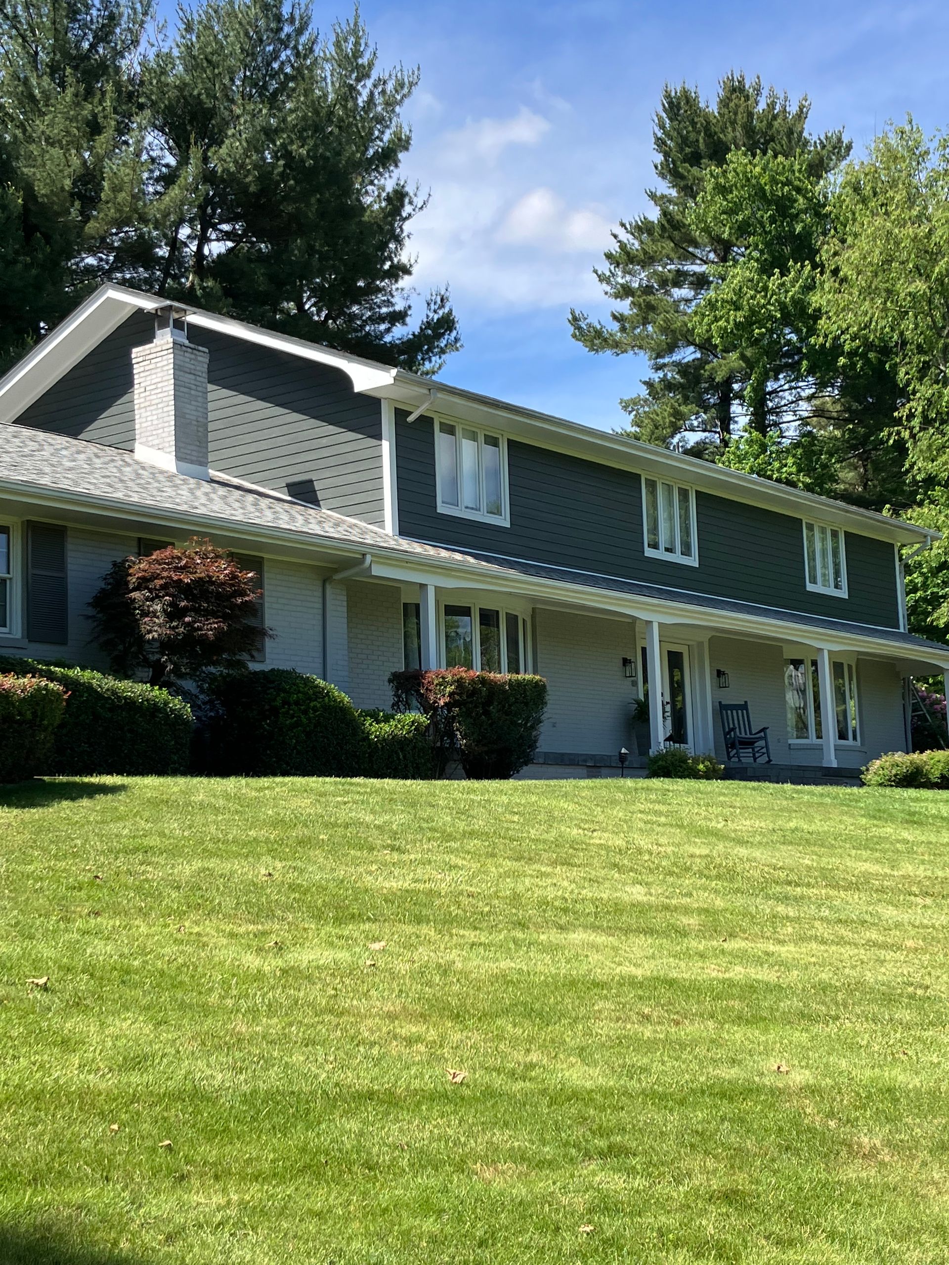A two-story house with dark gray siding and white trim, surrounded by trees and a green lawn under a sunny blue sky.