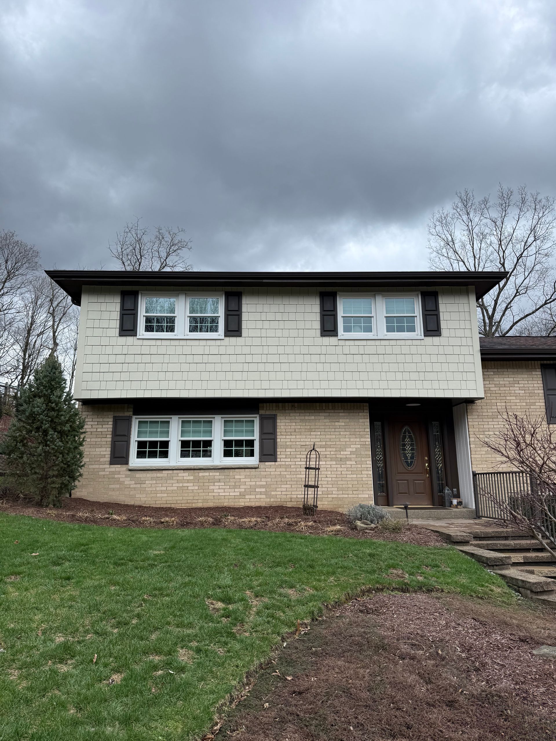 A two-story split-level home with beige siding on the upper level and stone brick on the lower level under a cloudy sky.