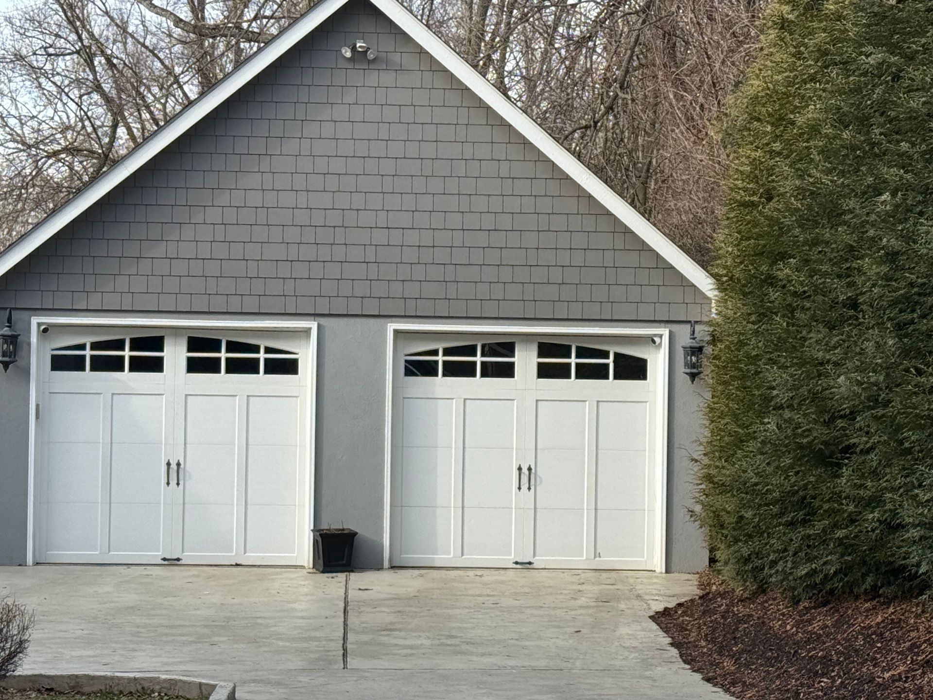 A two-car garage with white carriage-style doors and grey shingle siding, situated outdoors near trees.
