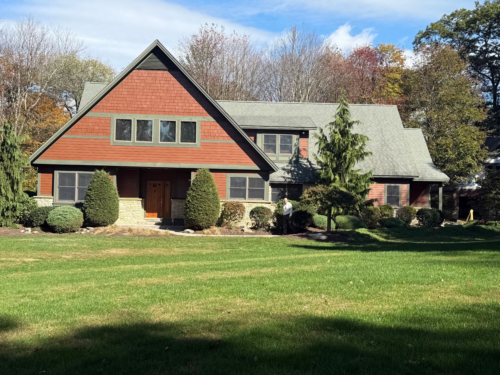 A red-sided house with a large triangular roof, set behind a green lawn surrounded by trees on a sunny day.