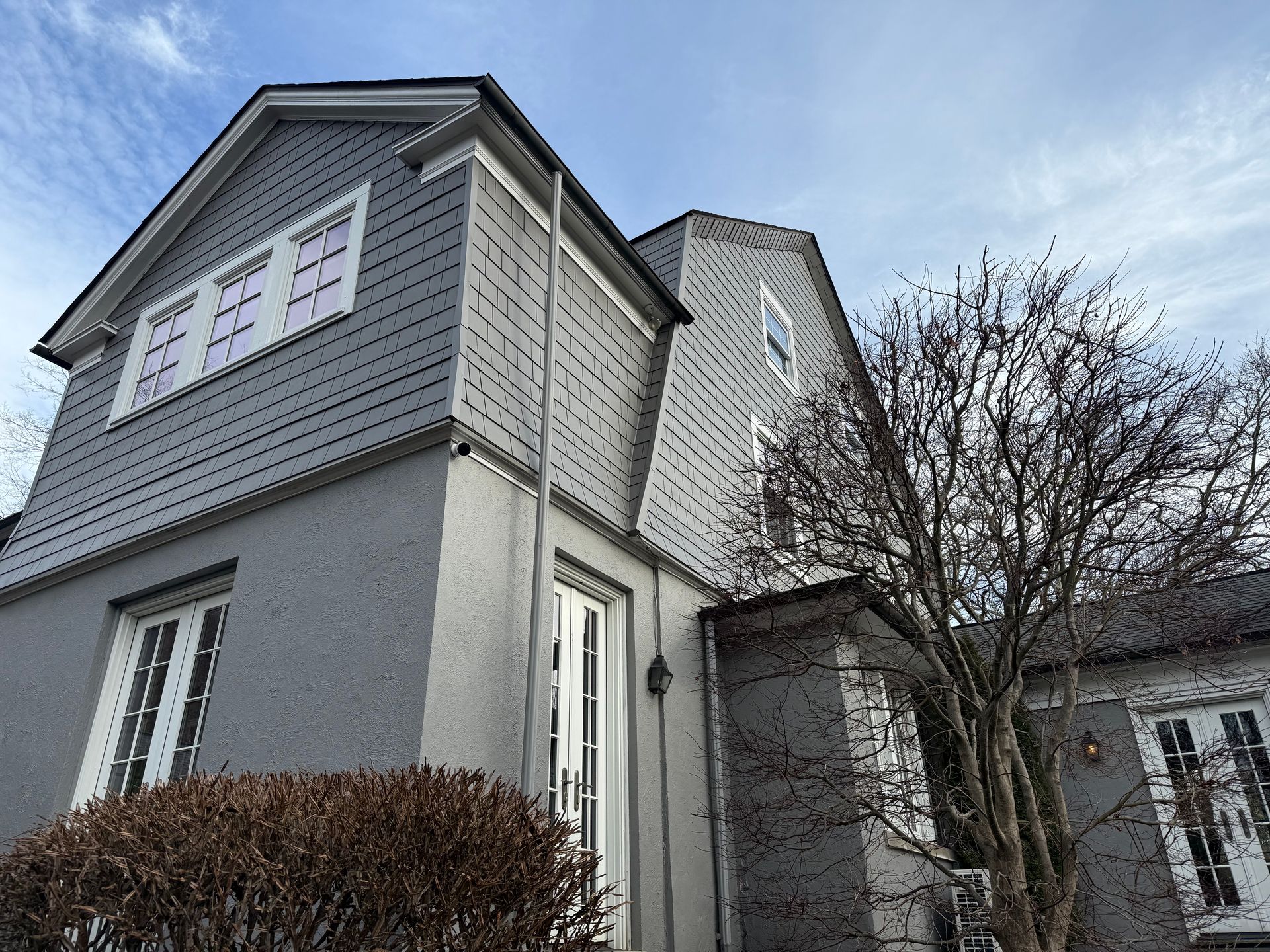 A low-angle view of a grey house with stucco lower walls, shingled upper stories, and multiple white-framed windows.