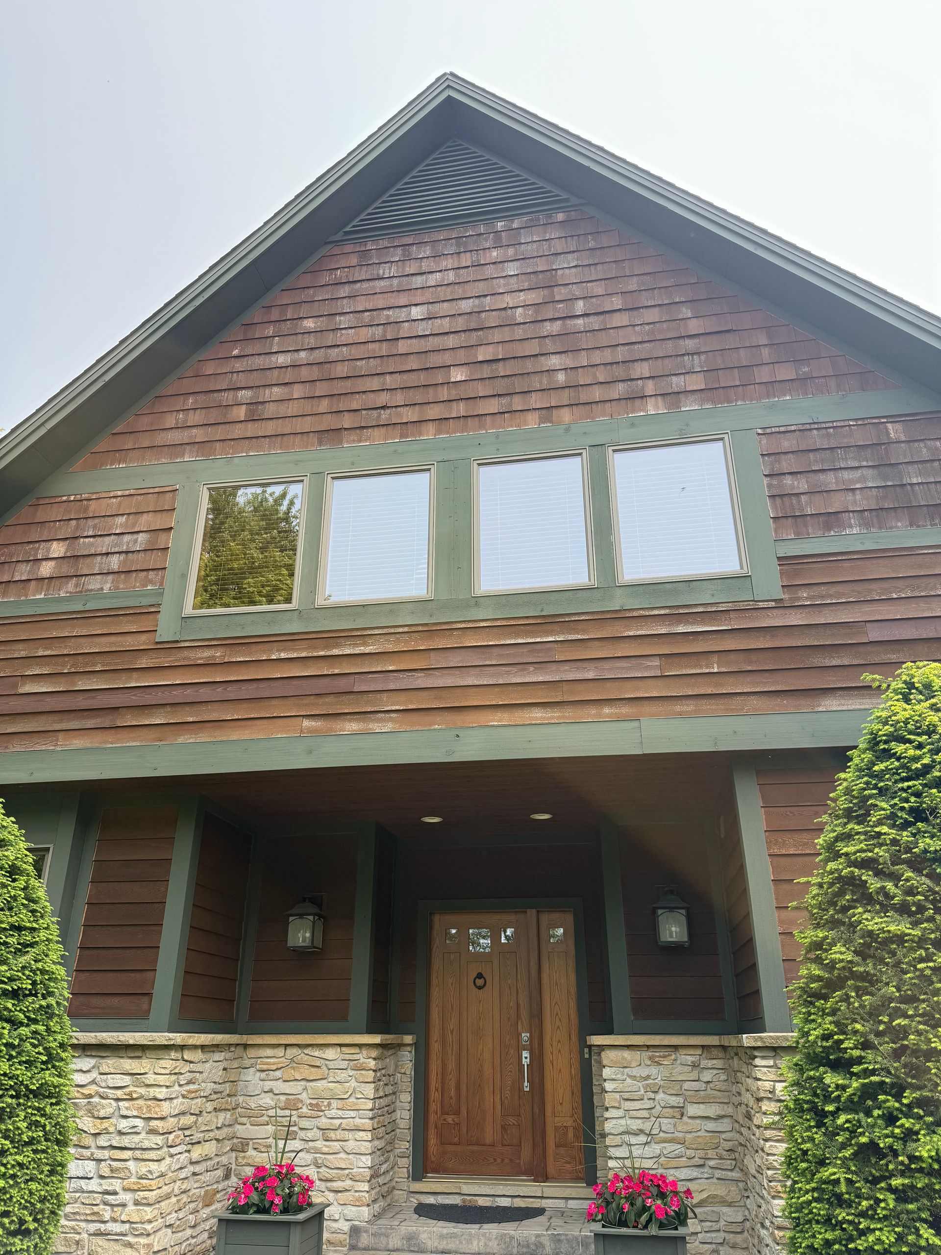 A brown wooden house with a stone base, a gabled roof, green trim, a central wooden door, and four rectangular windows.