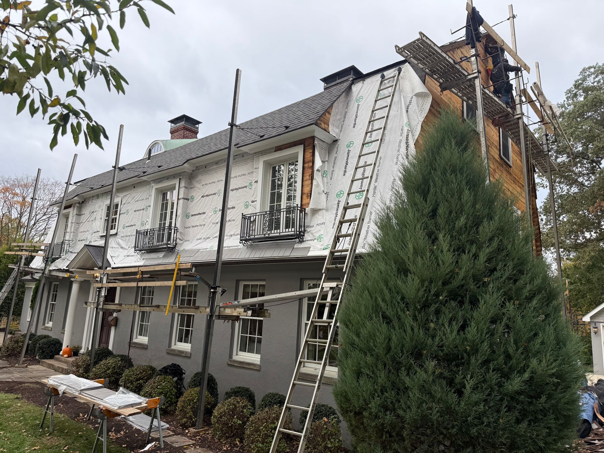 A two-story house under renovation, featuring exposed white weather barrier, scaffolding, and a ladder against the side.