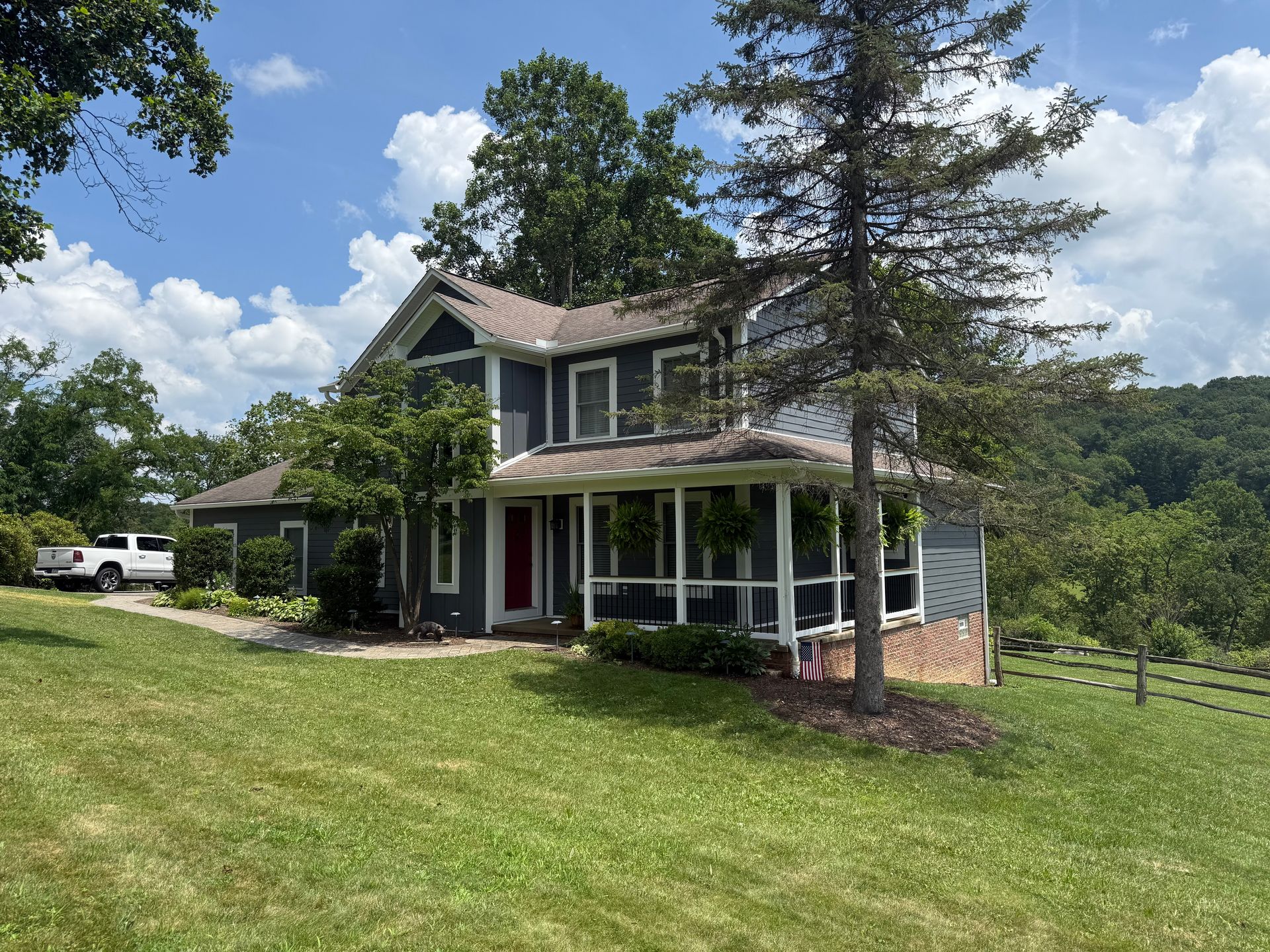 A two-story grey farmhouse with a white wrap-around porch, nestled in a grassy lawn with trees under a blue sky.