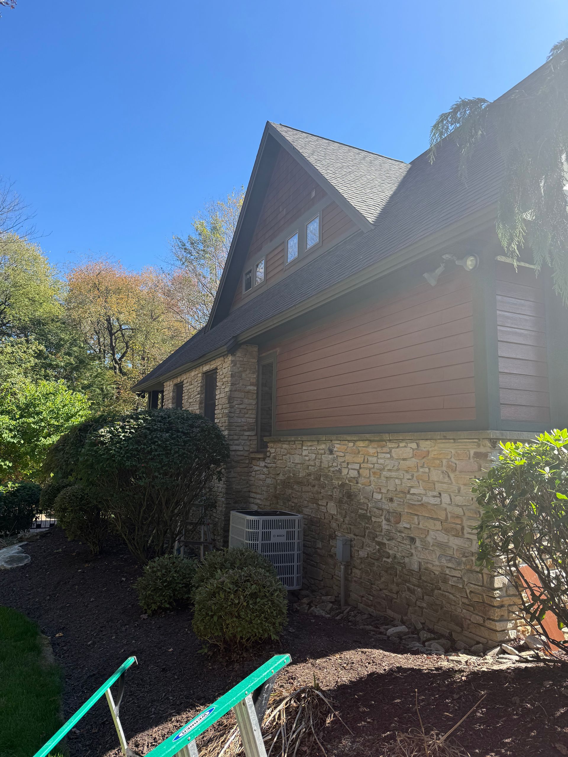 A side view of a house with a stone foundation, wood-shingled upper walls, and a peaked roof under a clear blue sky.