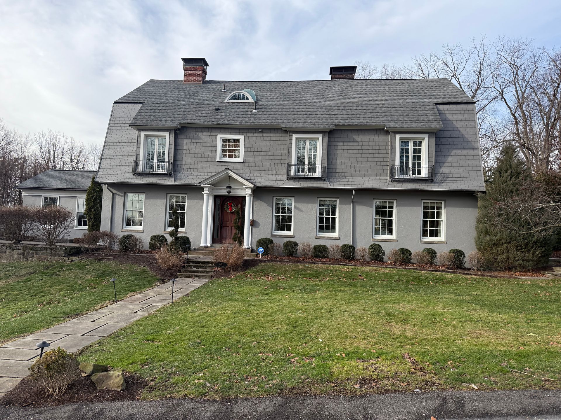 A large, two-story grey colonial-style house with a slate roof, three dormer windows, and a central front entrance.