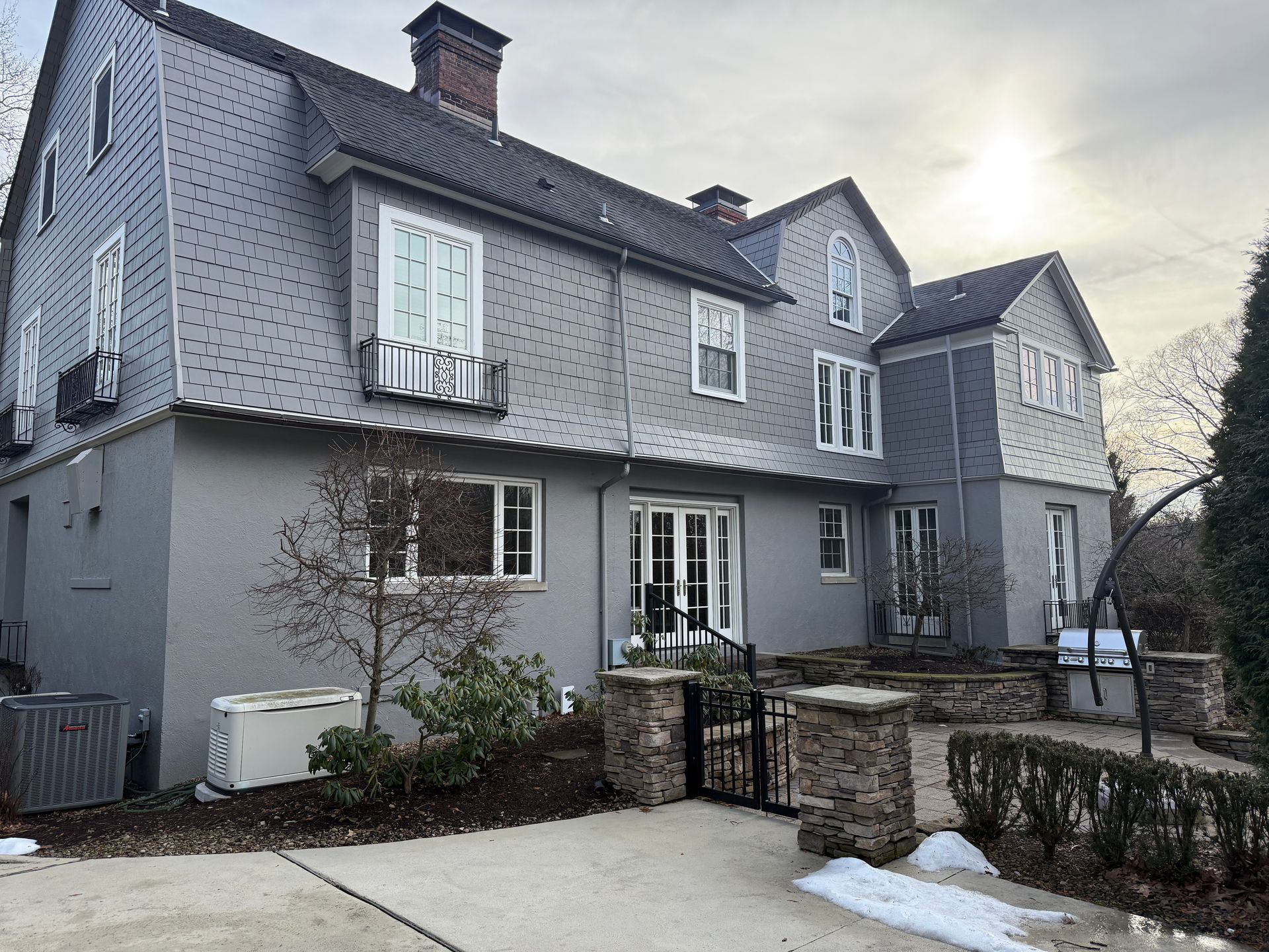 A large, two-story house with gray shingles on the upper floor, gray stucco on the lower, and a stone patio area.