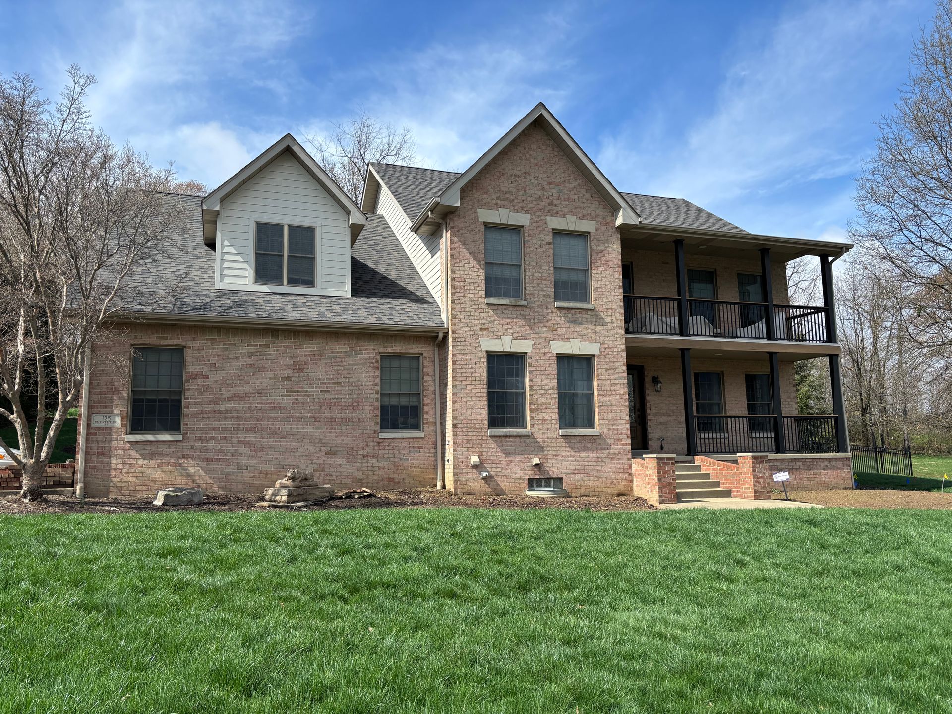 A two-story tan brick house with a dark shingled roof, a dormer, and a two-story covered porch, set on a green lawn.