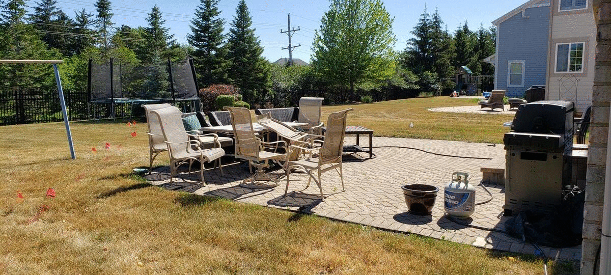 Backyard with patio furniture, a grill, and a trampoline, surrounded by a lawn and trees under a blue sky.