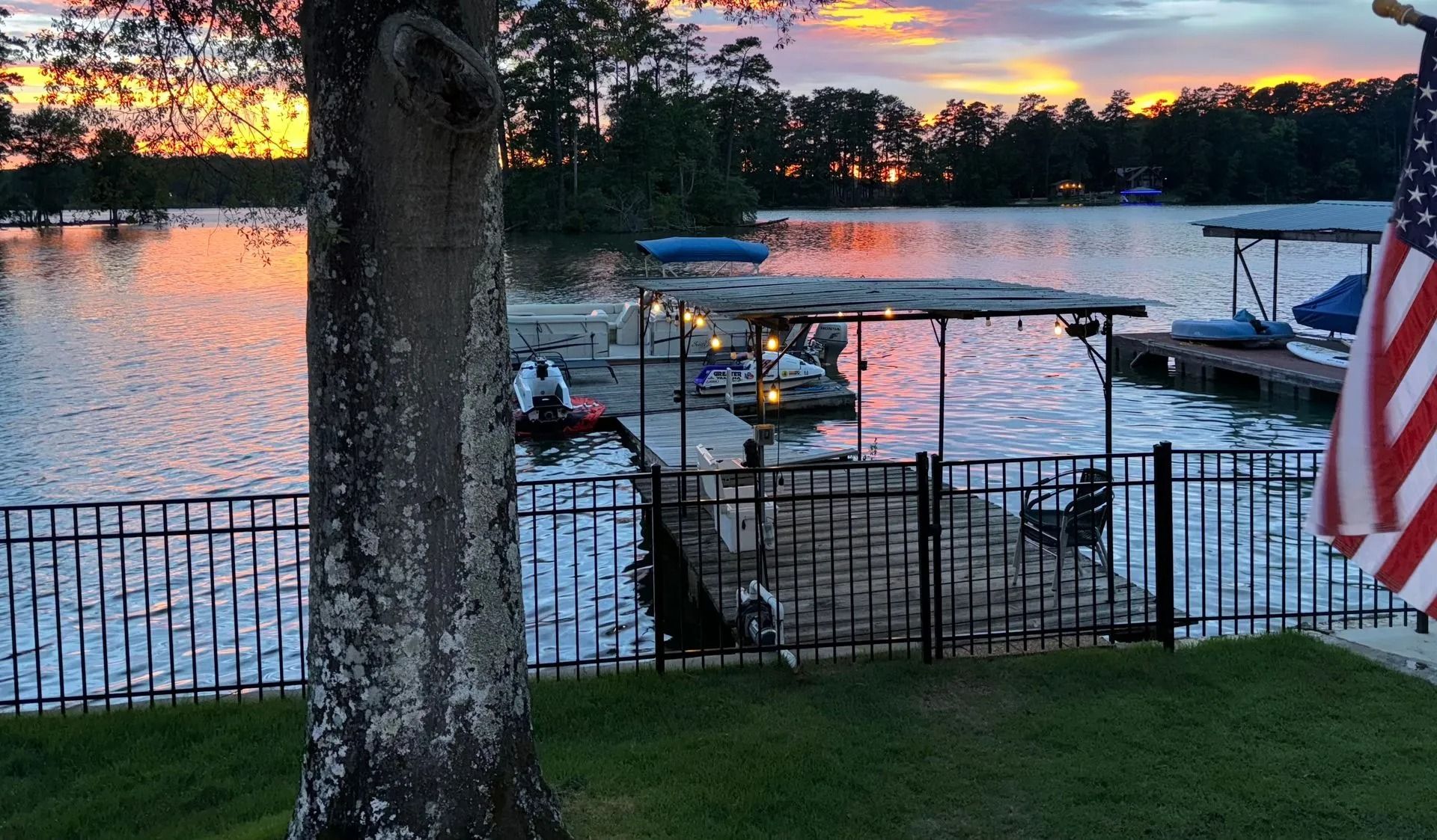 Sunset over a lakeside dock with boats, a fence, and an American flag in the foreground