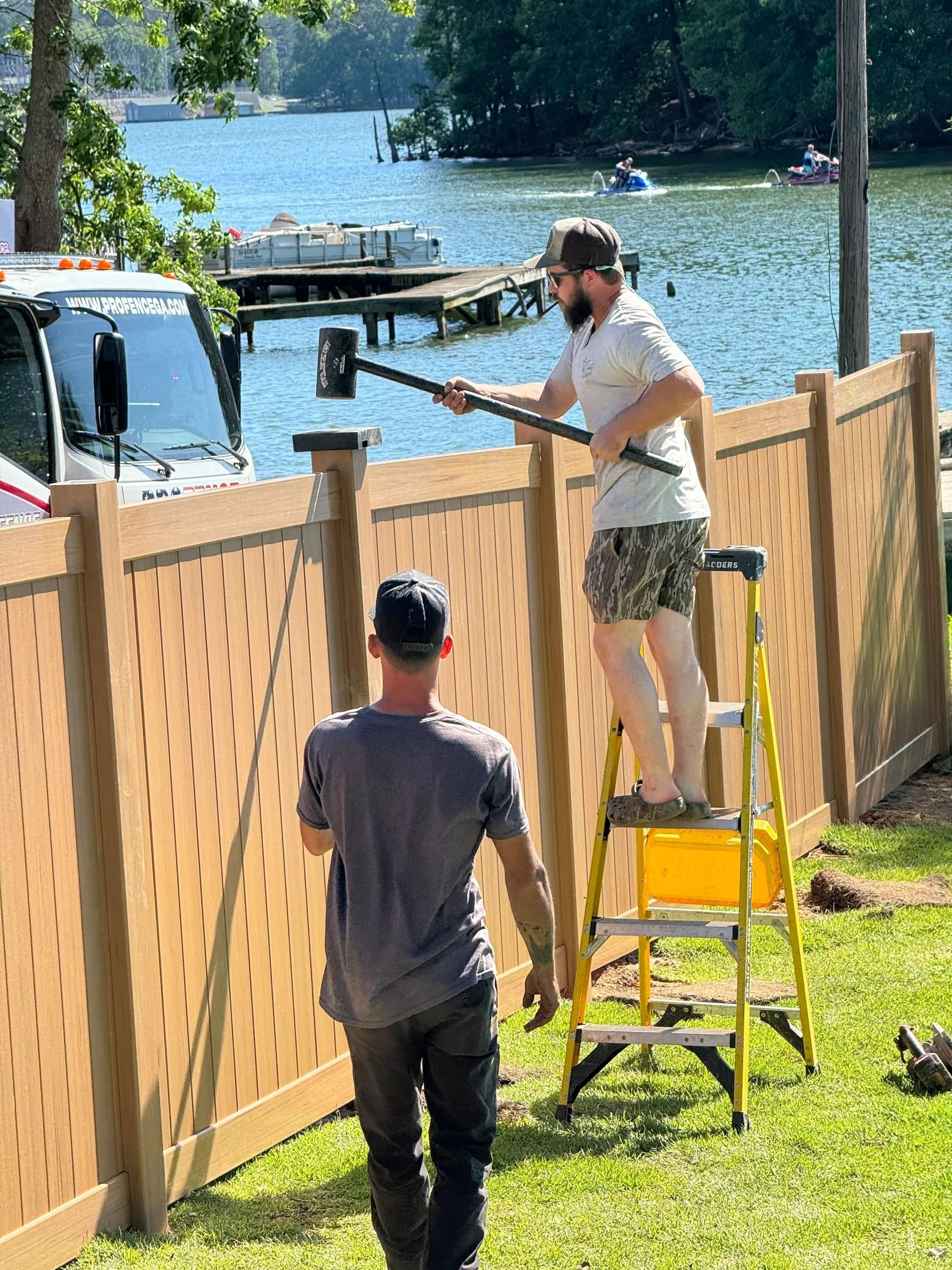 Two people installing a wooden fence by a lakeside, one standing on a yellow step ladder.