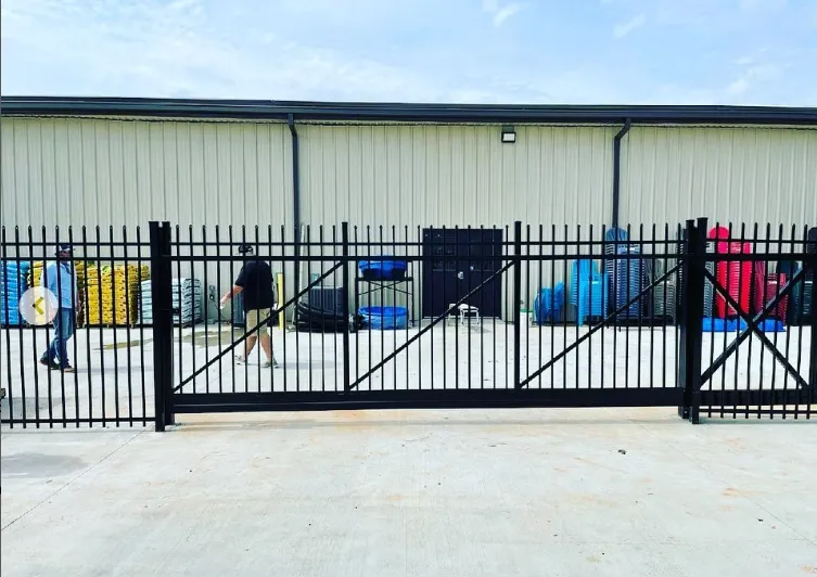 Black metal fence in front of a light industrial building with trash bins and a few people behind it