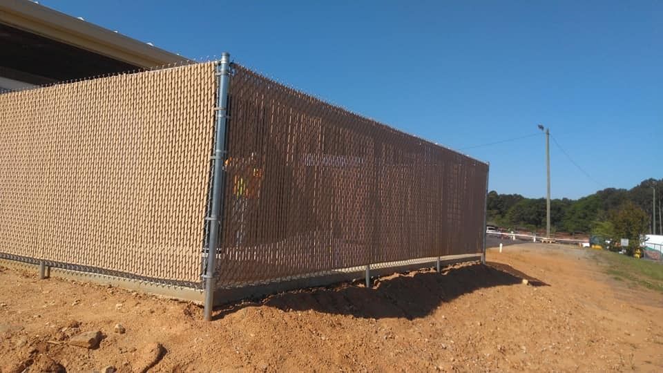 Chain-link fence around a dirt lot under a clear blue sky, with road and trees in the background.