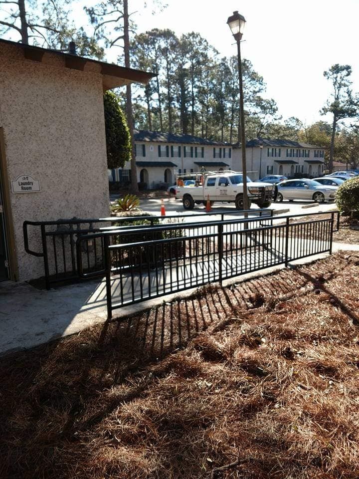Accessible ramp with black metal railings beside a building, leading toward a parking lot and apartment buildings.