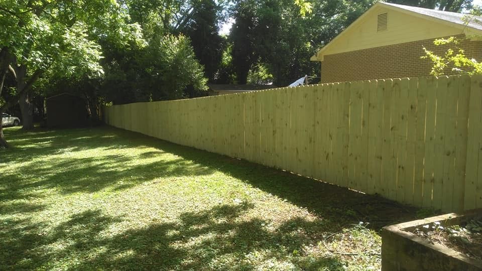 Sunny backyard with a long wooden fence, grass lawn, and a yellow house in the background