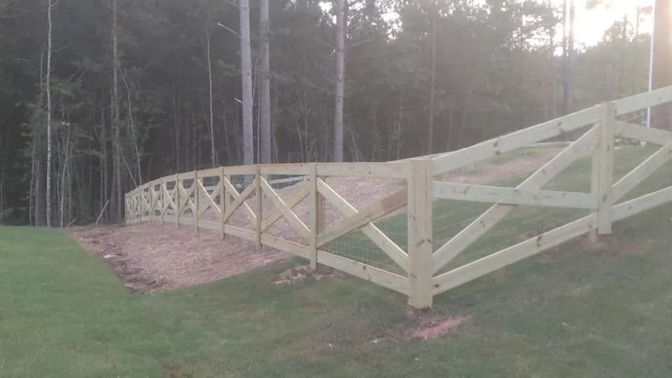 Wooden split-rail fence along a grassy yard beside a wooded area at dusk.