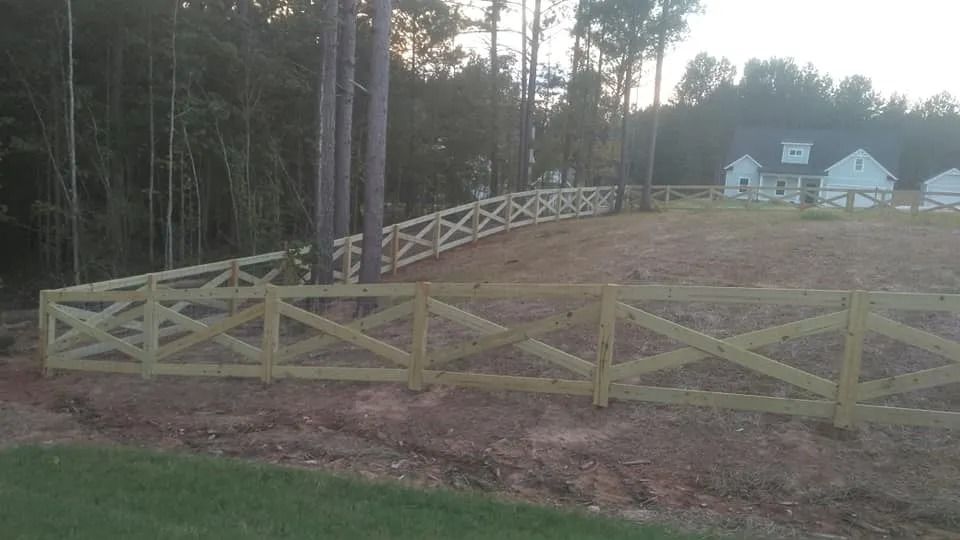 Wooden split-rail fence bordering a sloped grassy yard beside trees and houses at dusk