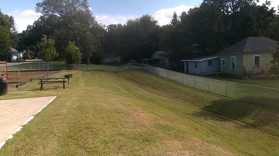 Grassy park field beside a road, with trees, a small building, and benches under a partly cloudy sky.