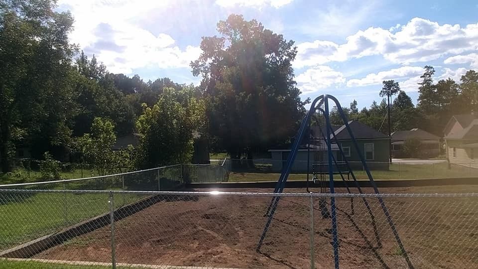 Chain-link fenced playground with blue climbing frame and dirt ground under sunny trees; houses in background