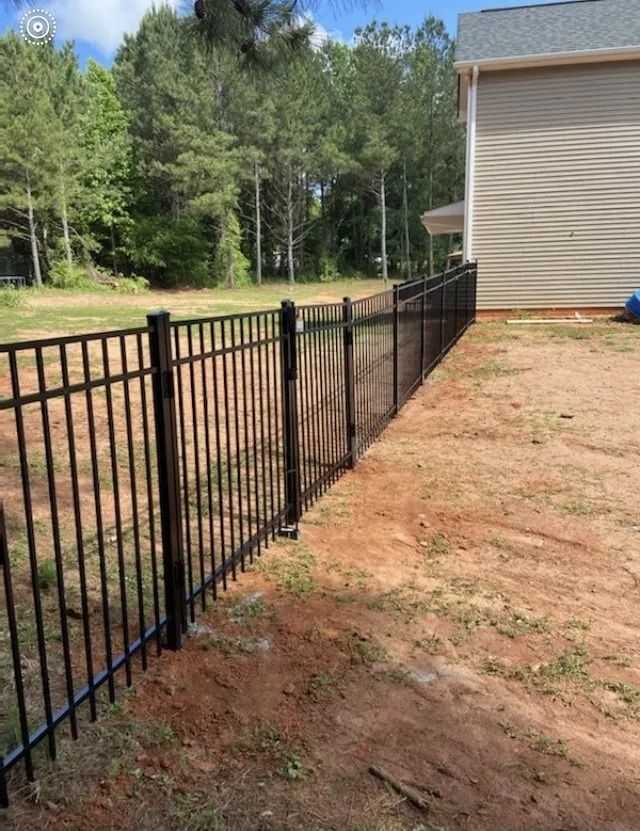 Black metal fence beside a tan house along a dirt yard with trees in the background