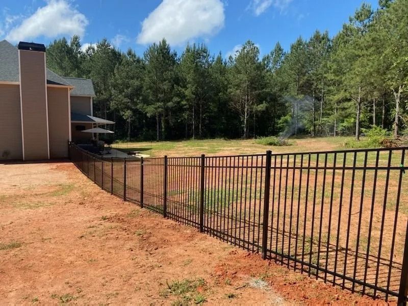 Black metal fence beside a tan building and red dirt yard, with trees and blue sky in the background