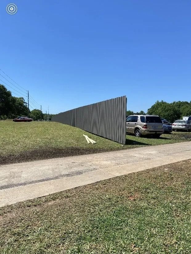 A grassy roadside by a sidewalk, with a long gray fence and parked vehicles under a clear blue sky.