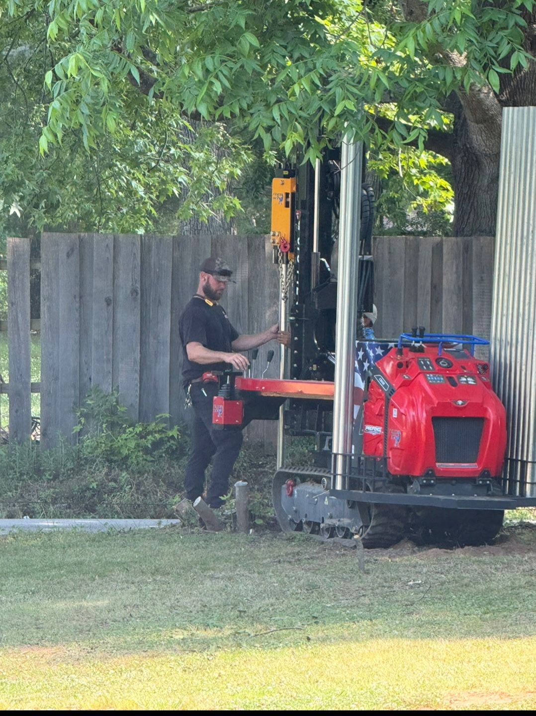 Worker operating a red utility machine beside a wooden fence and trees in a grassy yard
