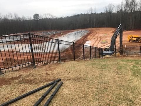 Construction site with graded dirt field, black fencing, and orange excavator near a wooded area