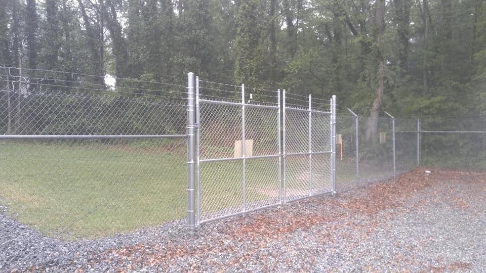Chain-link fence gate beside a gravel path, with trees and grassy field in the background.