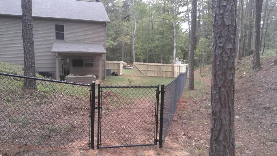 Open black gate in a wooded yard beside a gray house and fenced area.