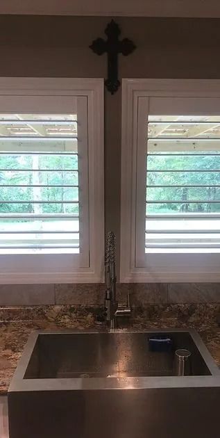 Kitchen sink beneath two windows with white shutters and a dark cross above.