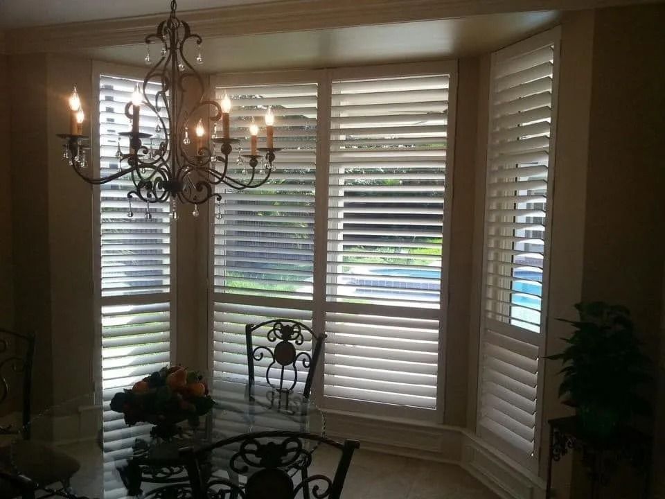 Sunlit dining room with bay windows, white blinds, and a dark chandelier over a glass table.