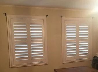 Two white plantation shutters on a beige wall with sunlight filtering through the slats.