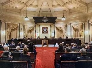 Courtroom with seated audience facing a panel under ornate ceiling and red carpet center aisle