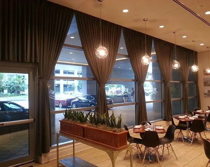 Modern café interior with tall windows, hanging lights, and small tables and chairs near a planter