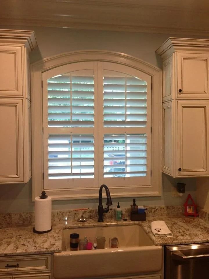 Kitchen sink beneath a window with white shutters, granite counters, and light wood cabinets.