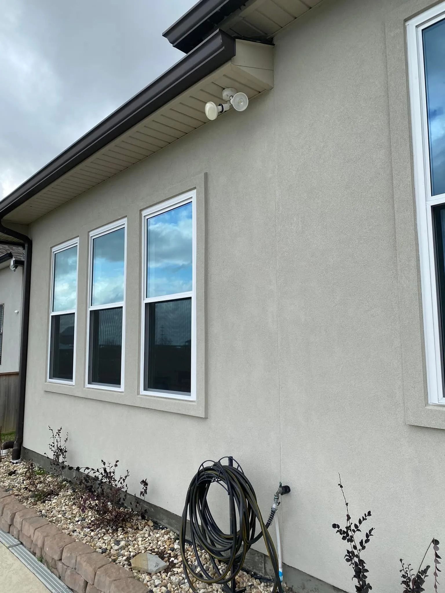 Exterior stucco wall with three windows, white trim, and coiled hose beneath the eaves